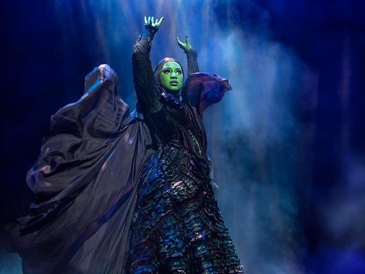 Dynamic scene from the musical Wicked, featuring Elphaba and Glinda performing on a lavishly decorated stage with dramatic lighting