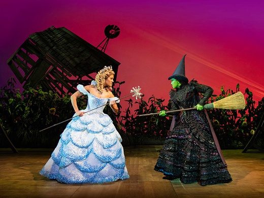 Dynamic scene from the musical Wicked, featuring Elphaba and Glinda performing on a lavishly decorated stage with dramatic lighting