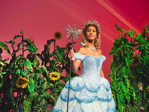 Dynamic scene from the musical Wicked, featuring Elphaba and Glinda performing on a lavishly decorated stage with dramatic lighting