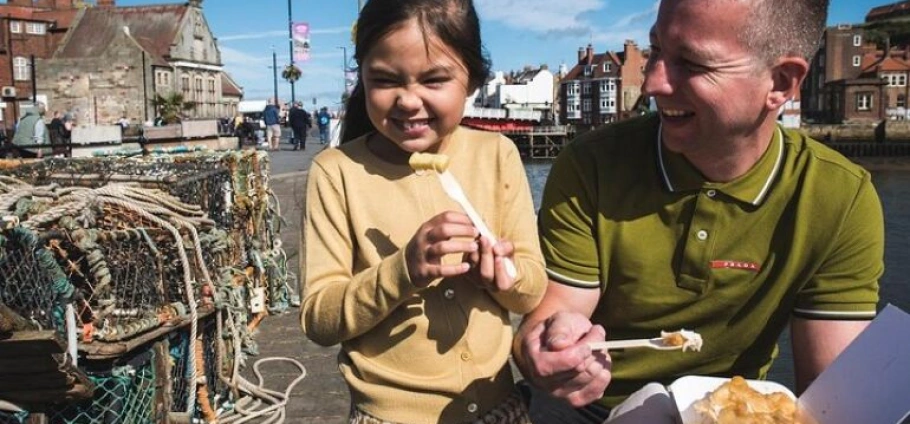 Father and daughter enjoying fish and chips by the harbor on a sunny day.