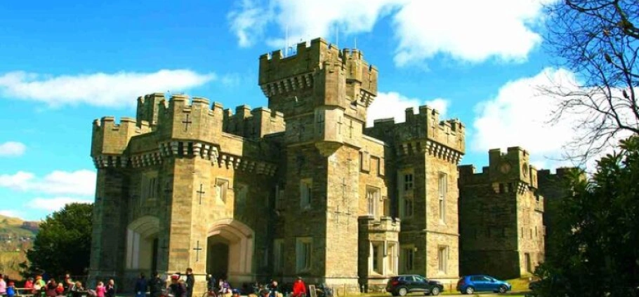 Historic stone castle with turrets and battlements under a blue sky, surrounded by visitors and parked cars.