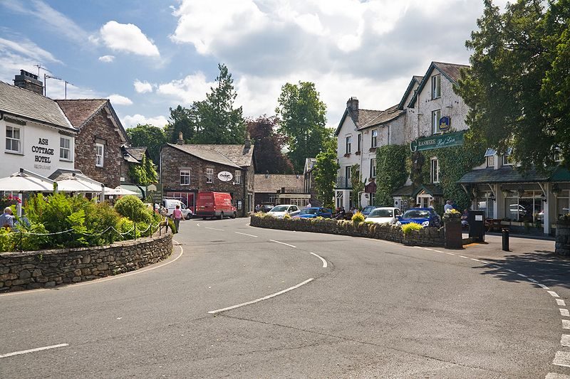 Charming village scene featuring Ash Cottage Hotel and surrounding buildings on a sunny day, with a curved road, parked cars, and lush greenery.