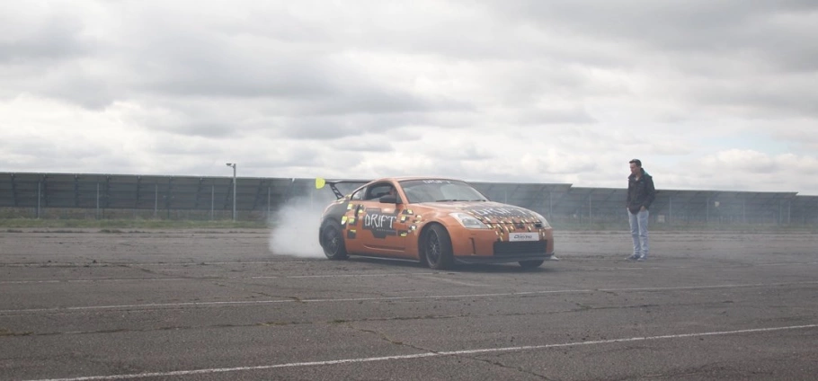 Orange drift car performing a burnout with smoke on an empty asphalt lot under cloudy skies, person standing nearby.