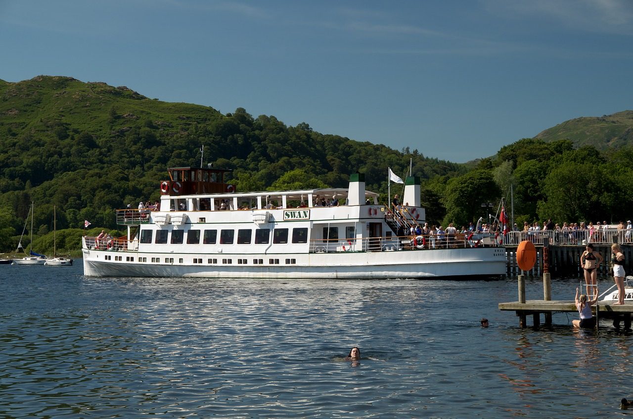 A white passenger boat named "Swan" navigates a serene lake on a sunny day, surrounded by lush green hills. People are seen enjoying the boat ride, while others swim and relax on a nearby dock.