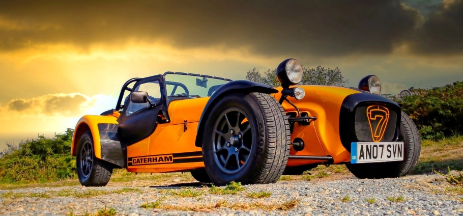 Orange Caterham 7 sports car parked on gravel road with dramatic sunset sky in the background.