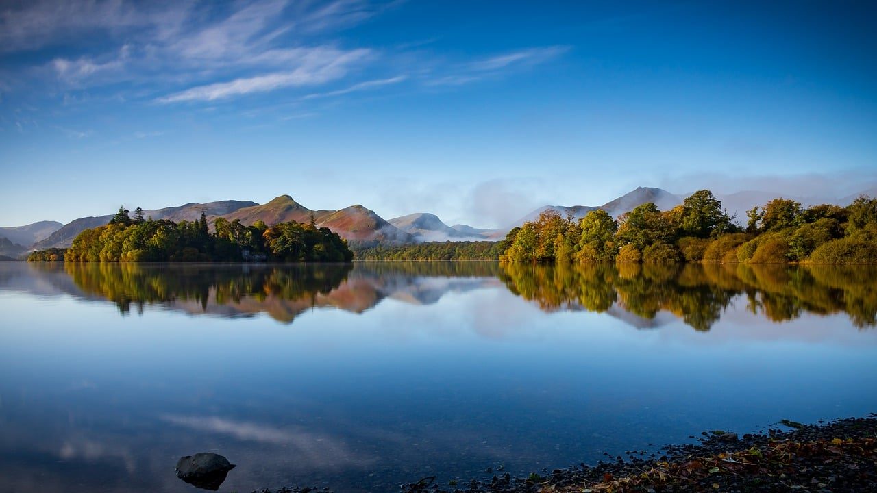 Scenic view of a tranquil lake surrounded by lush greenery and rolling hills, reflecting a clear blue sky and distant mountains, ideal for nature lovers and outdoor enthusiasts.