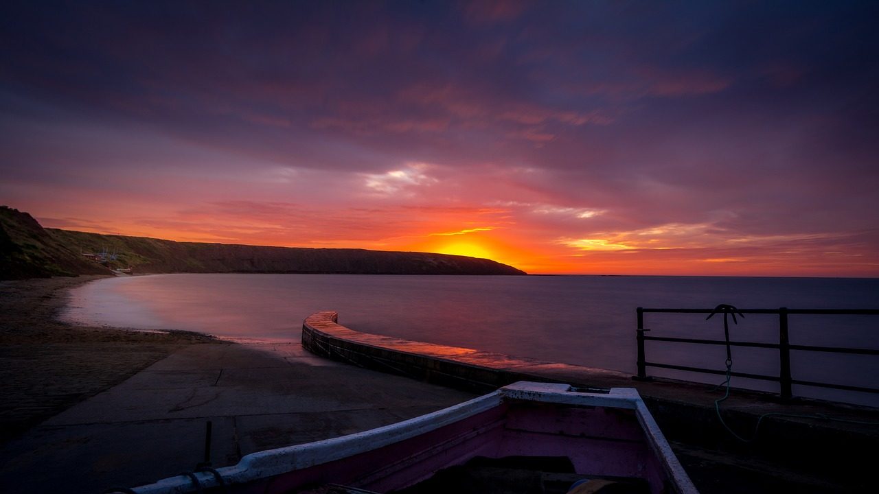 Scenic sunset over a calm beach with a curved pier, showcasing vibrant orange and purple hues in the sky, and a partially visible boat in the foreground.