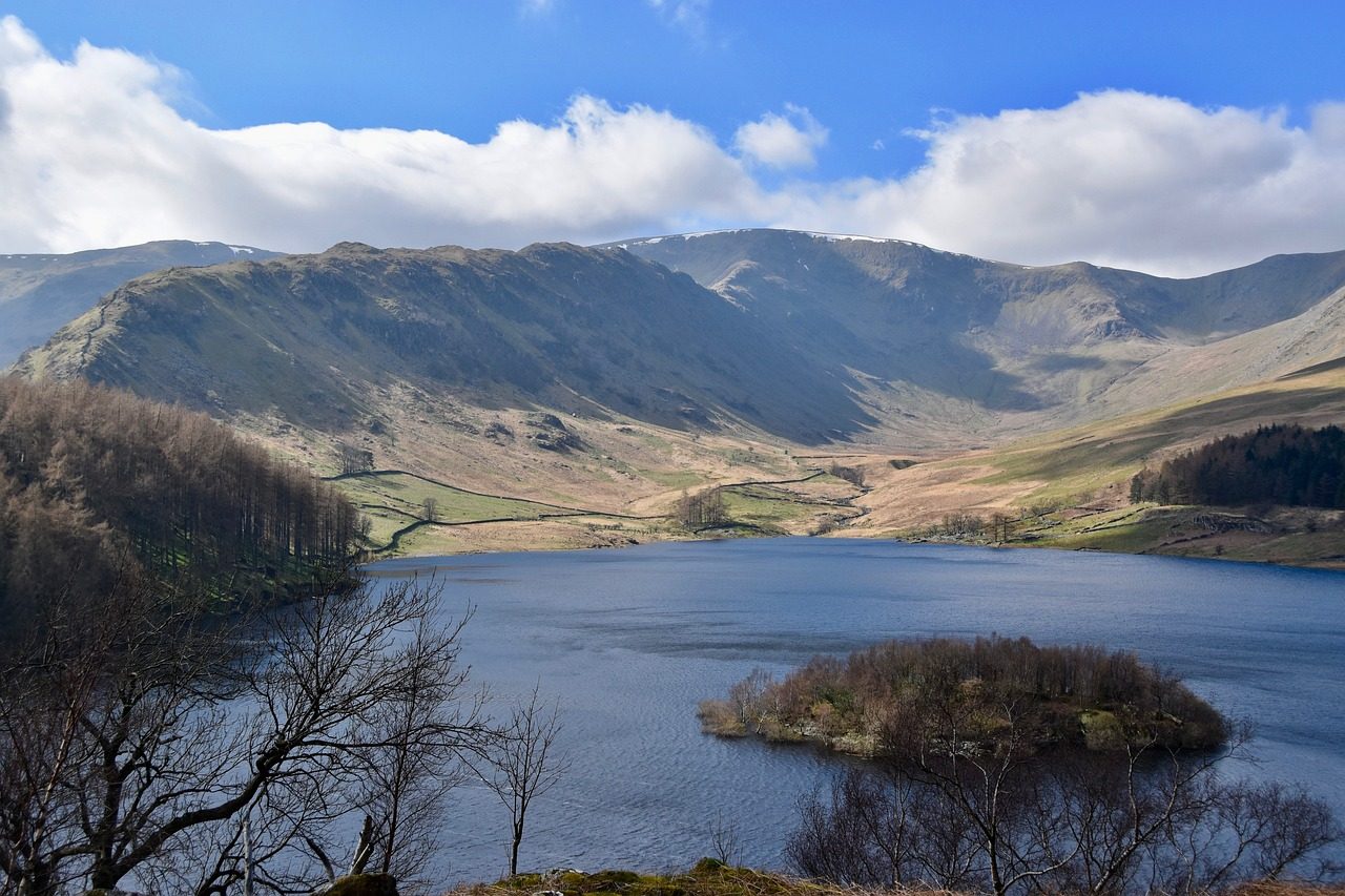 Scenic view of a tranquil lake surrounded by rolling hills and mountains under a partly cloudy sky, showcasing the natural beauty of the landscape.