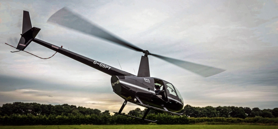 Black helicopter flying low over a grassy field with trees in the background.