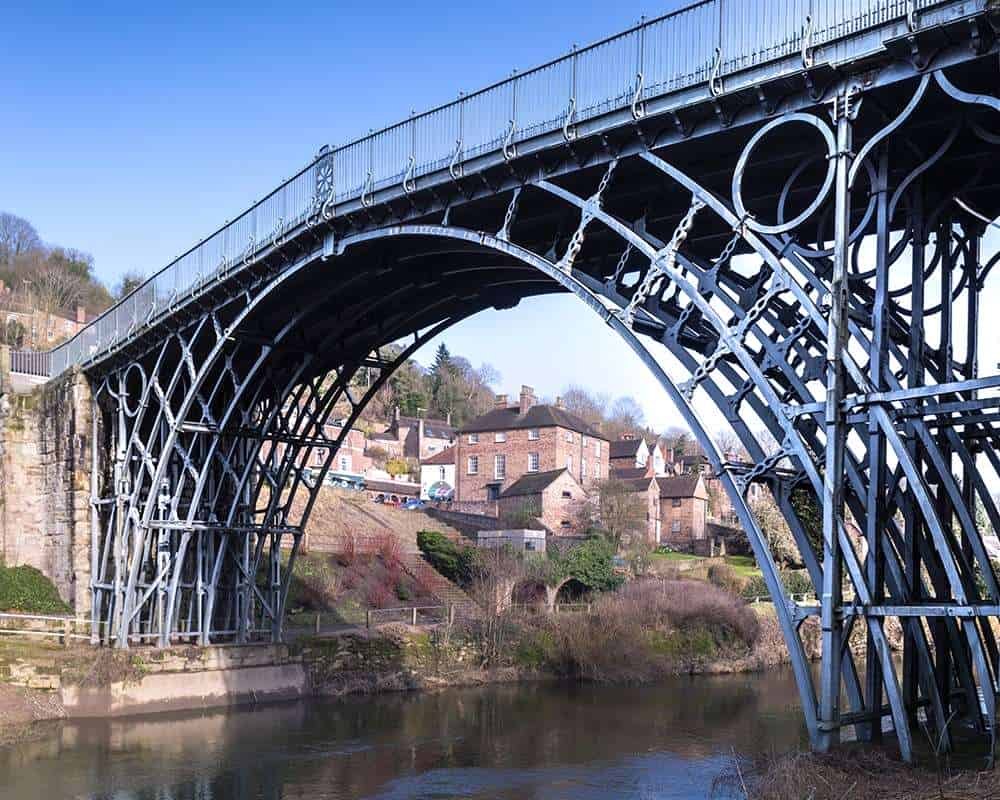 Close-up view of the cast-iron arch structure of the historic Iron Bridge crossing the River Severn, with town buildings visible underneath.