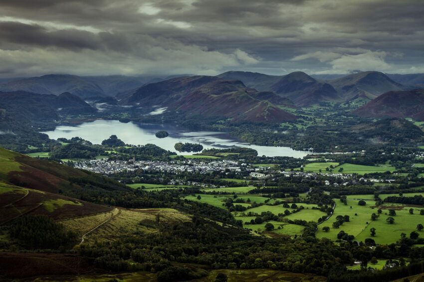 Keswick in the lake district | Aerial view of a lush green landscape featuring a serene lake surrounded by mountains, with a small town visible near the water's edge under a cloudy sky.