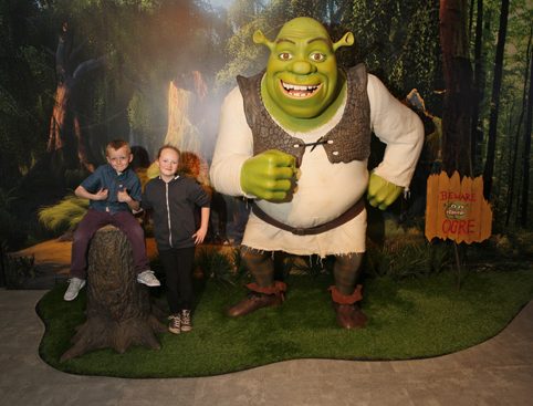 Two children posing with a life-sized Shrek statue in a forest-themed setting