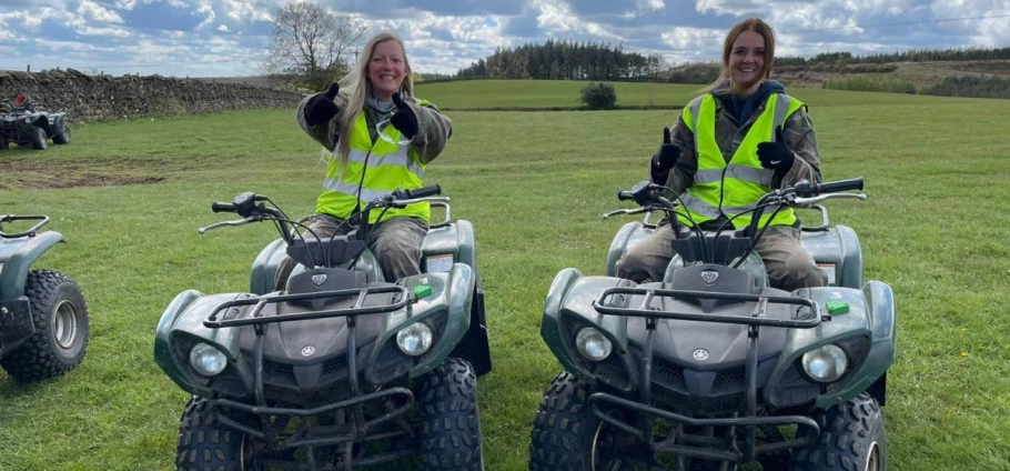 Two women in high-visibility vests riding ATVs on a grassy field with a scenic countryside background.