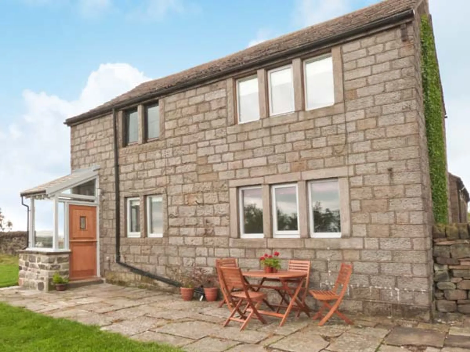 Knowle Lodge | Stone cottage with a wooden door and large windows, featuring a patio area with a small table and chairs, surrounded by greenery and a scenic landscape. Ideal for outdoor relaxation and enjoying nature.