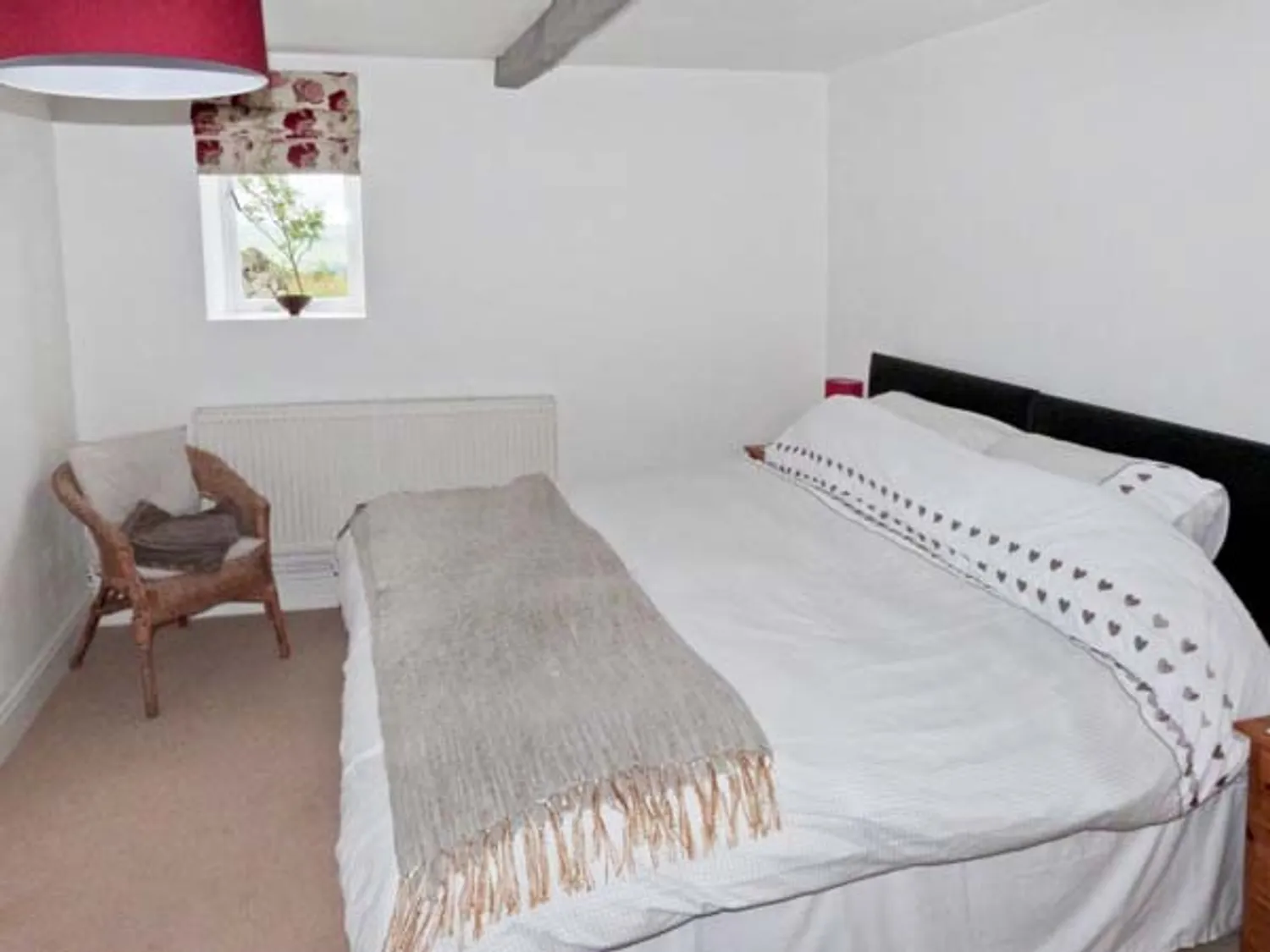 Cozy bedroom featuring a large bed with white bedding and decorative throw, a wicker chair, and a window adorned with a floral curtain, creating a warm and inviting atmosphere.