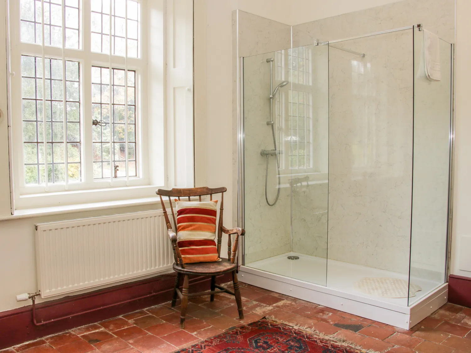 Spacious bathroom with glass shower enclosure, vintage wooden chair with striped cushion, large window with garden view, and terracotta tiled floor.