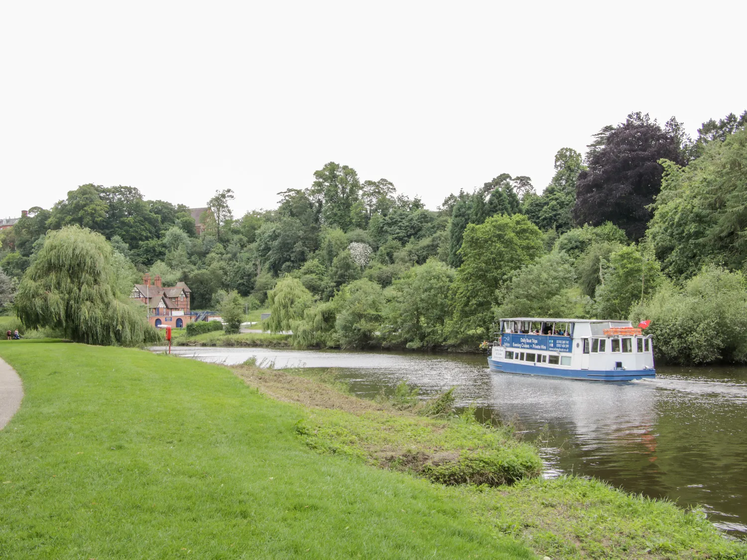 Scenic river with a tour boat cruising past lush green trees and a charming riverside cottage.