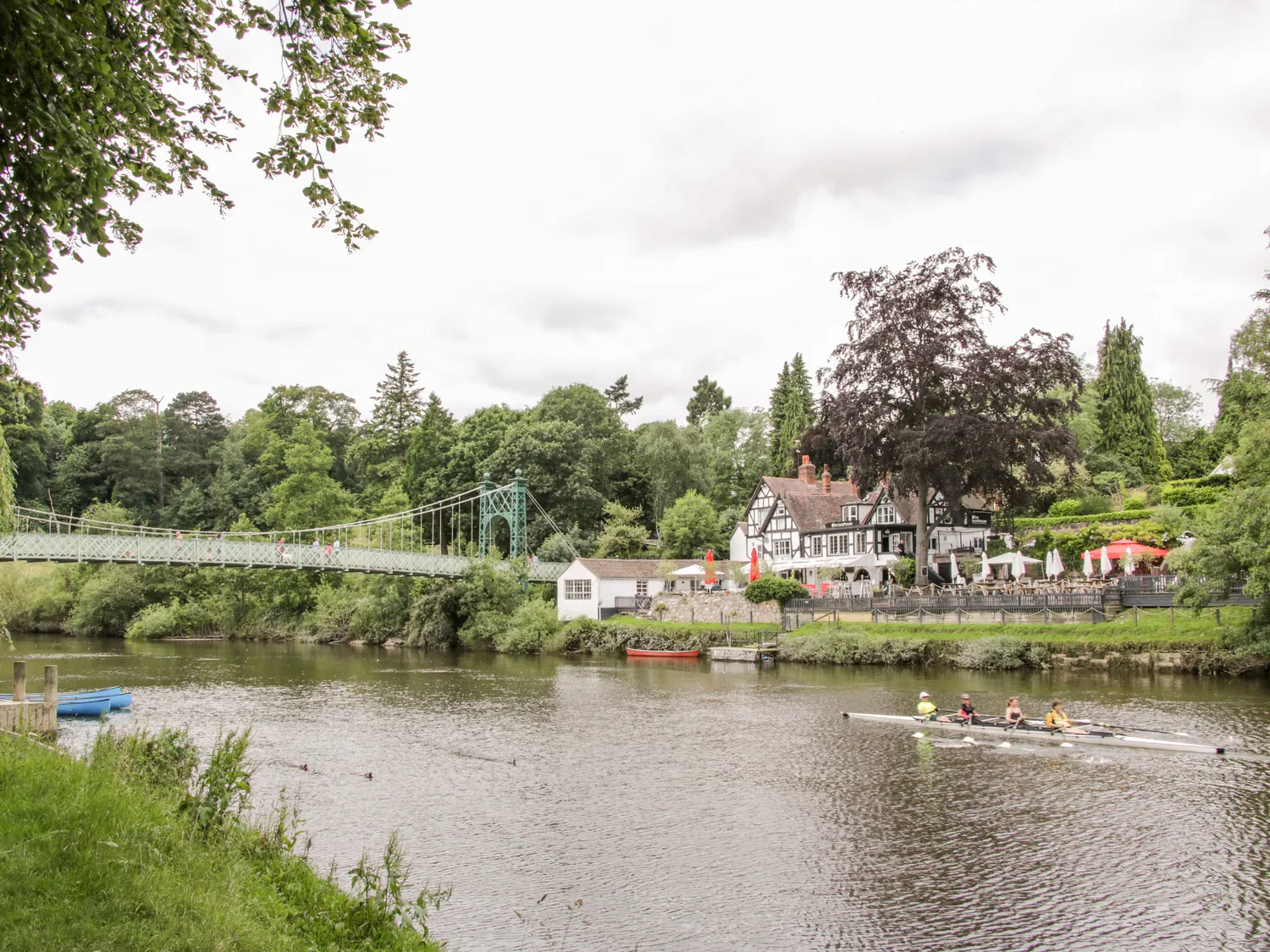 Scenic riverside view with a suspension bridge, traditional house, and people rowing on the river.