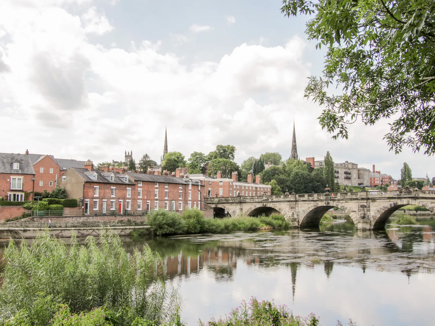 Historic town with brick houses and stone bridge over a river, lush greenery and church spires in the background.