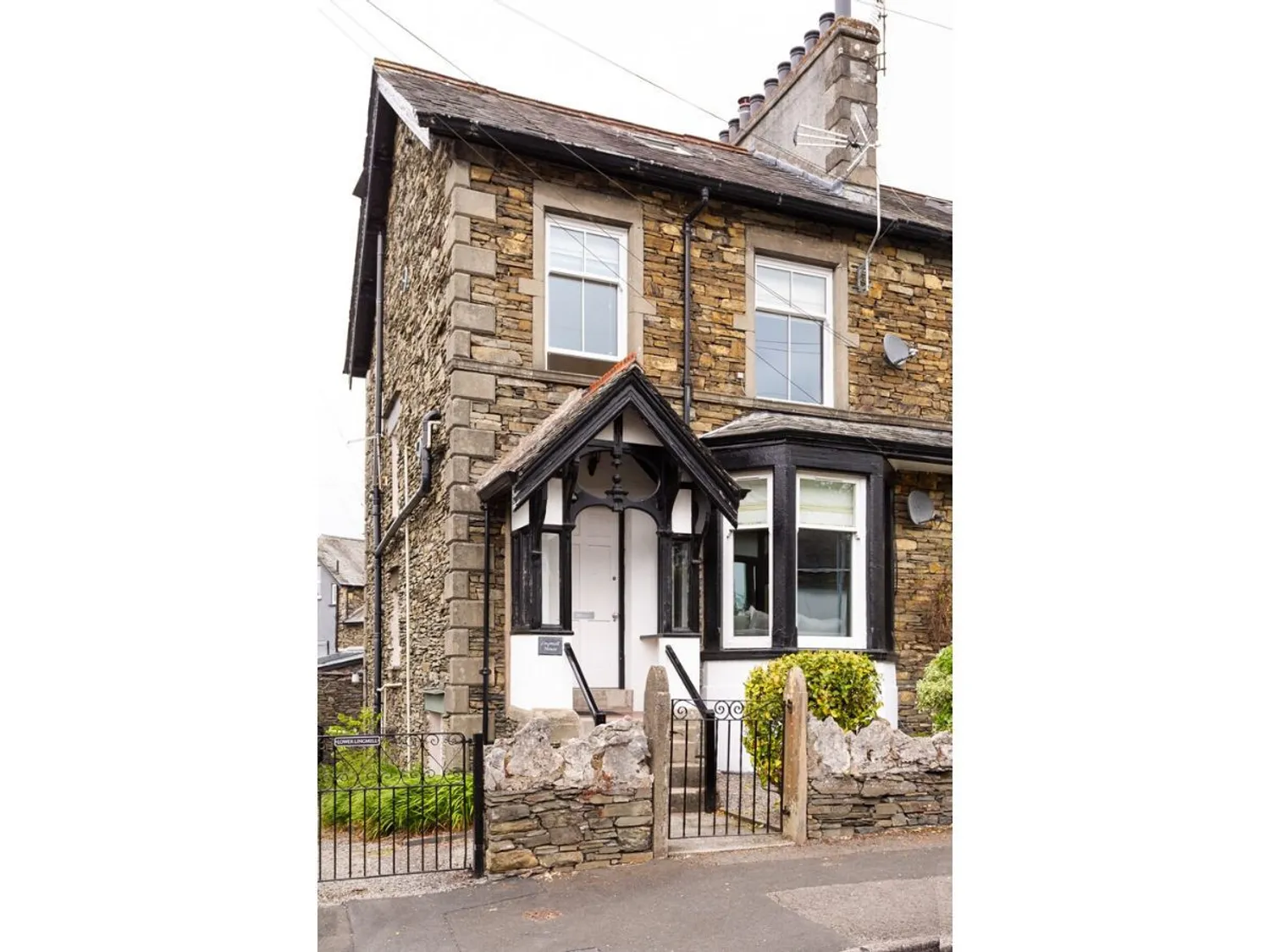 Lingmell House in Bowness | Victorian-style stone house with a black and white entrance, featuring a wrought iron gate and manicured shrubs, located on a quiet street.