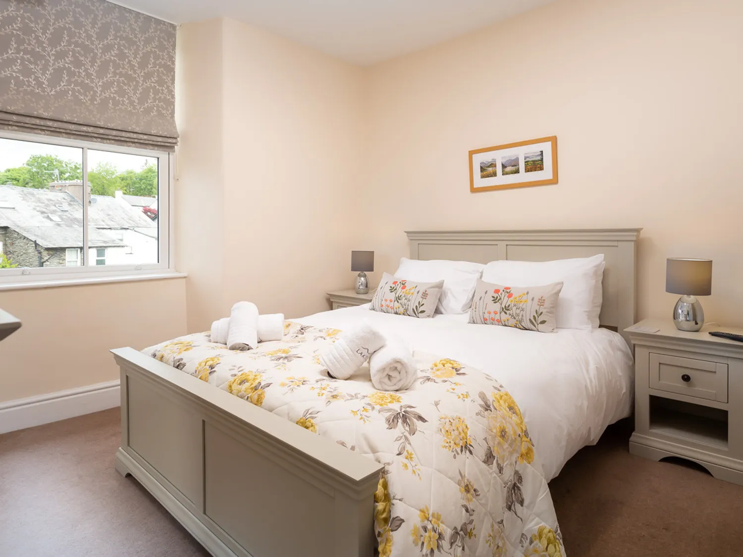 Cozy bedroom featuring a queen-sized bed with floral bedding, decorative pillows, and neatly arranged towels. The room includes bedside tables with lamps and a window offering natural light and views of the outdoors. Ideal for a relaxing stay.