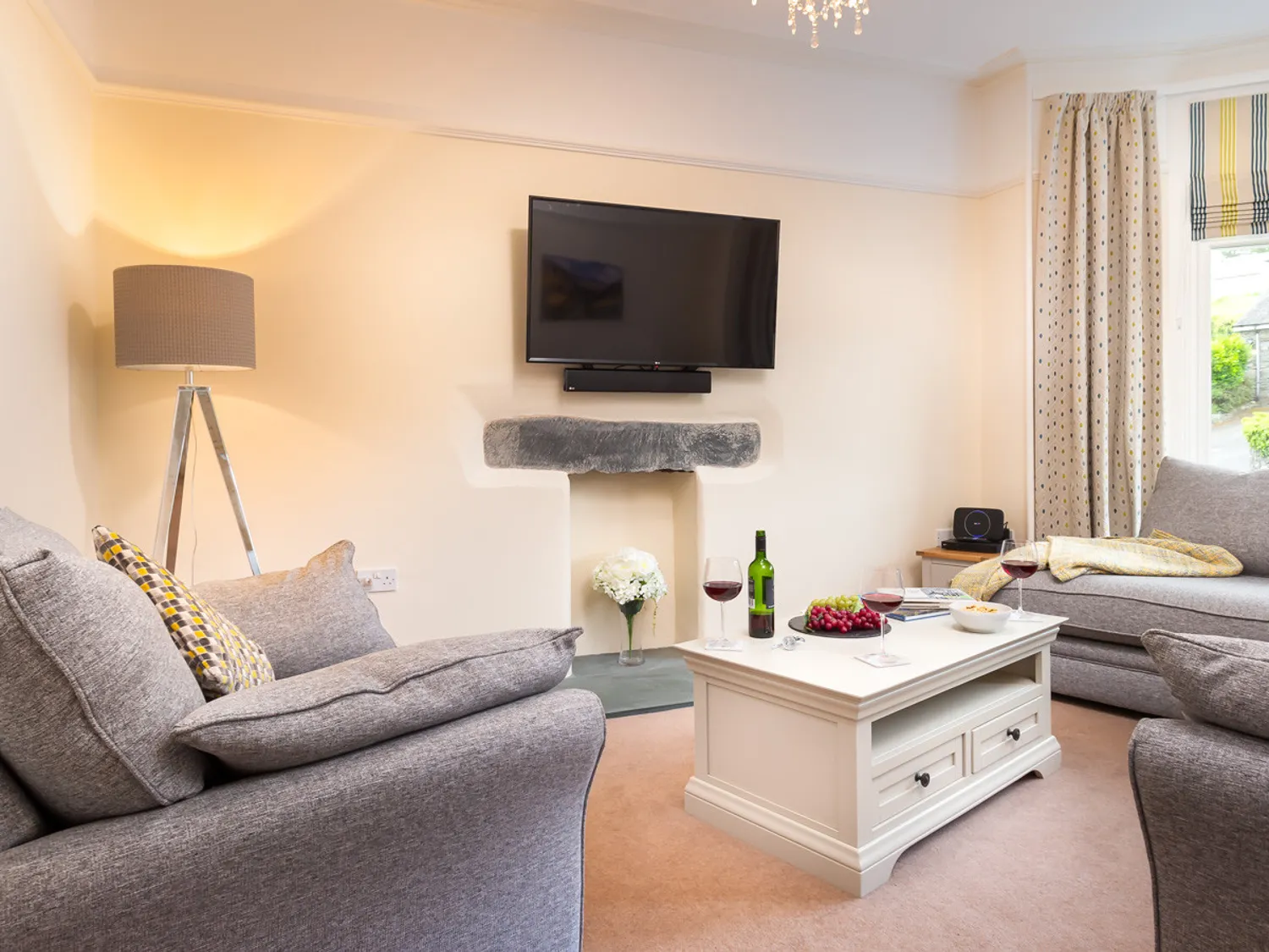 Cozy living room featuring a gray sofa, a stylish coffee table with wine glasses, and a flat-screen TV mounted above a stone fireplace. Soft lighting from a modern lamp and patterned curtains enhance the inviting atmosphere.