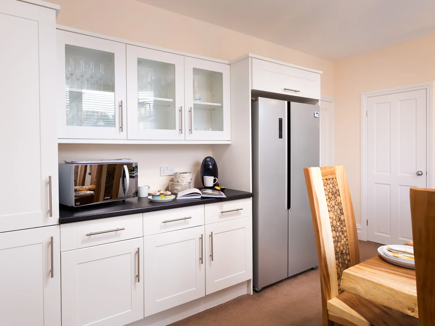 Modern kitchen interior featuring white cabinetry, stainless steel appliances, and a wooden dining table. The kitchen includes a microwave, coffee maker, and decorative tableware, creating a stylish and functional space.