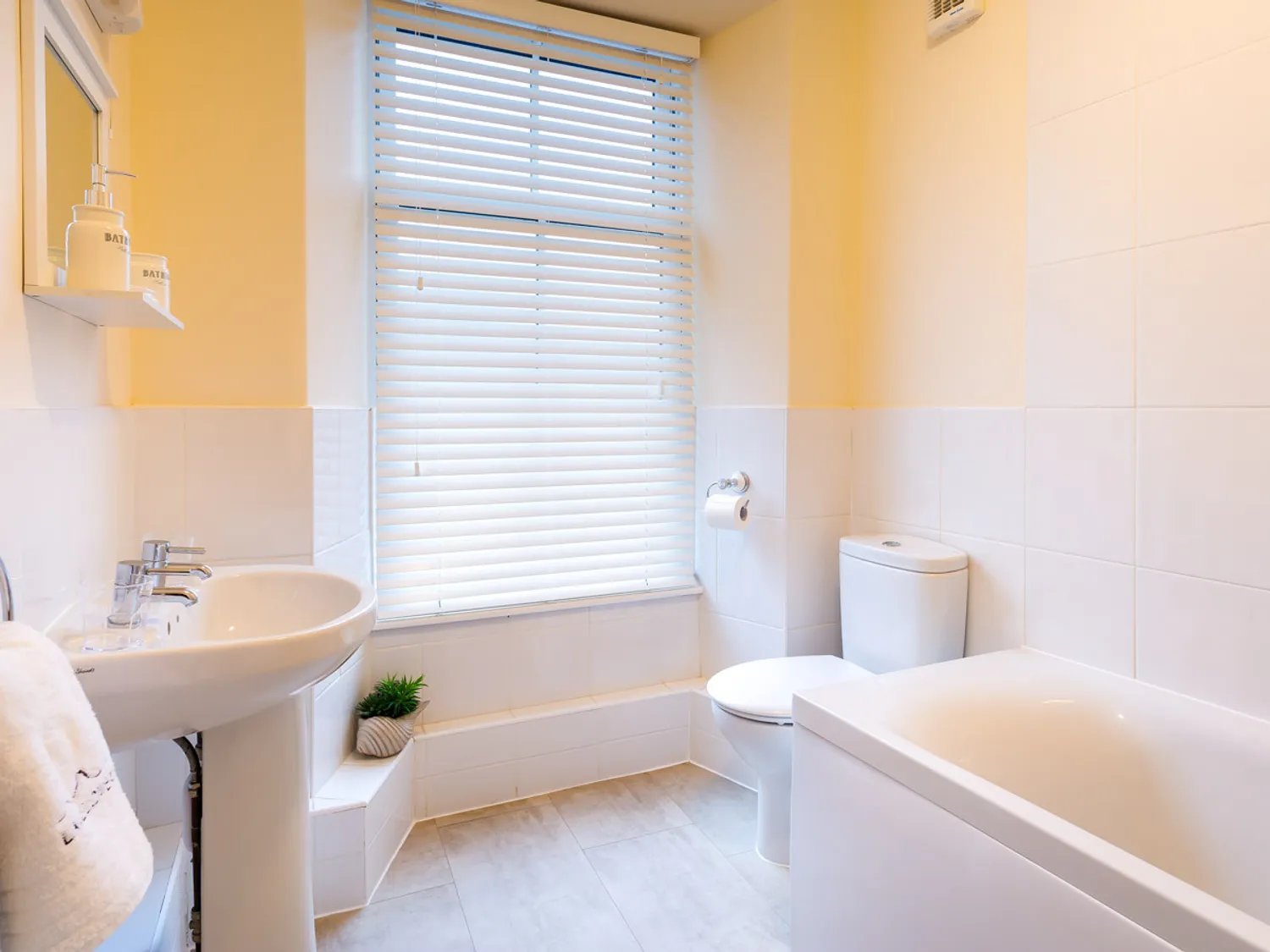 Bright and modern bathroom featuring a white sink, toilet, and bathtub, with natural light streaming in through a window adorned with blinds. The decor includes a small plant and neatly arranged toiletries, creating a clean and inviting atmosphere.