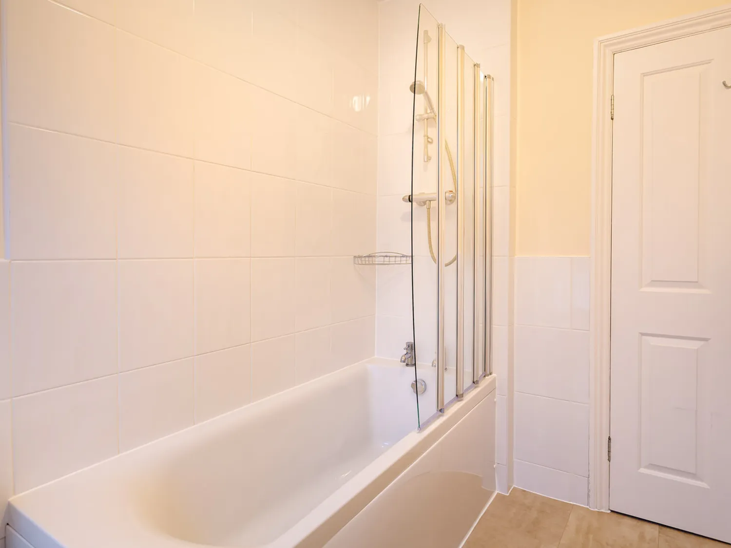 Modern bathroom featuring a white bathtub with a glass shower screen, light-colored tiled walls, and a door leading to another space. The design emphasizes cleanliness and functionality.