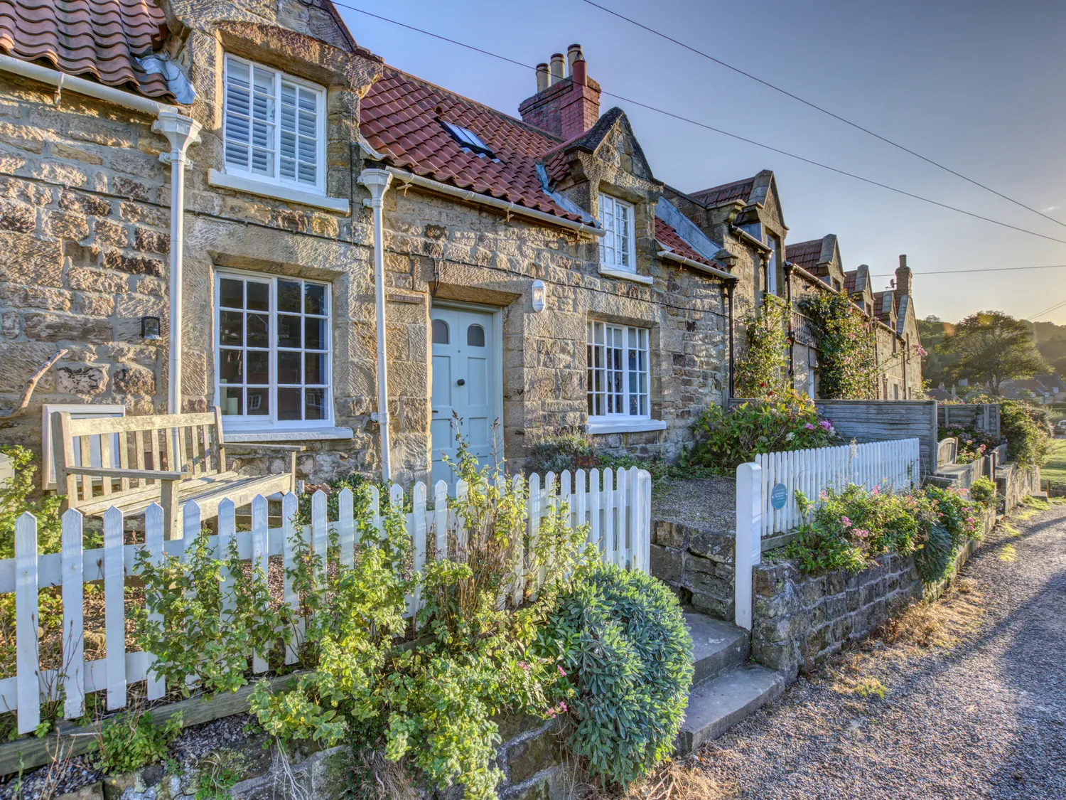 Charming stone cottage with white picket fence and red tiled roof in a picturesque countryside setting.