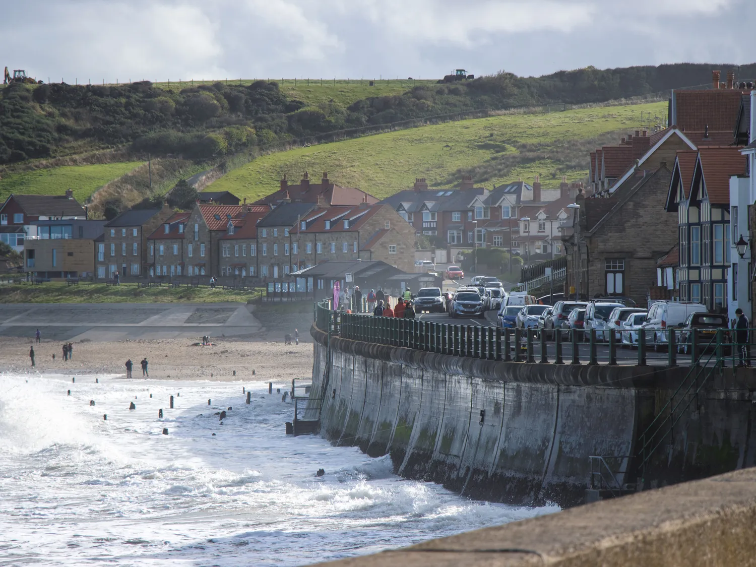 Coastal town with seaside promenade, parked cars, and people walking along the beach on a cloudy day.