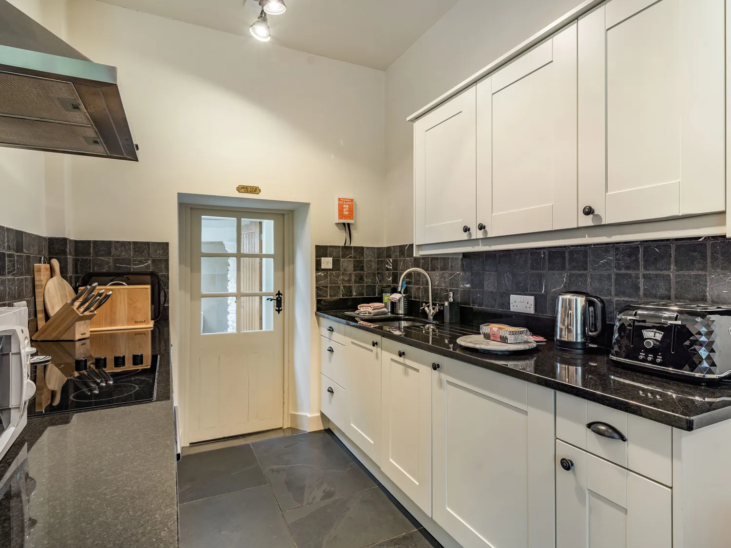 Coachman's Cottage: Modern kitchen featuring sleek black countertops, white cabinetry, and stainless steel appliances, including a kettle and toaster. The space has a rustic wooden cutting board and a glass door leading outside, complemented by stylish dark tiled walls.