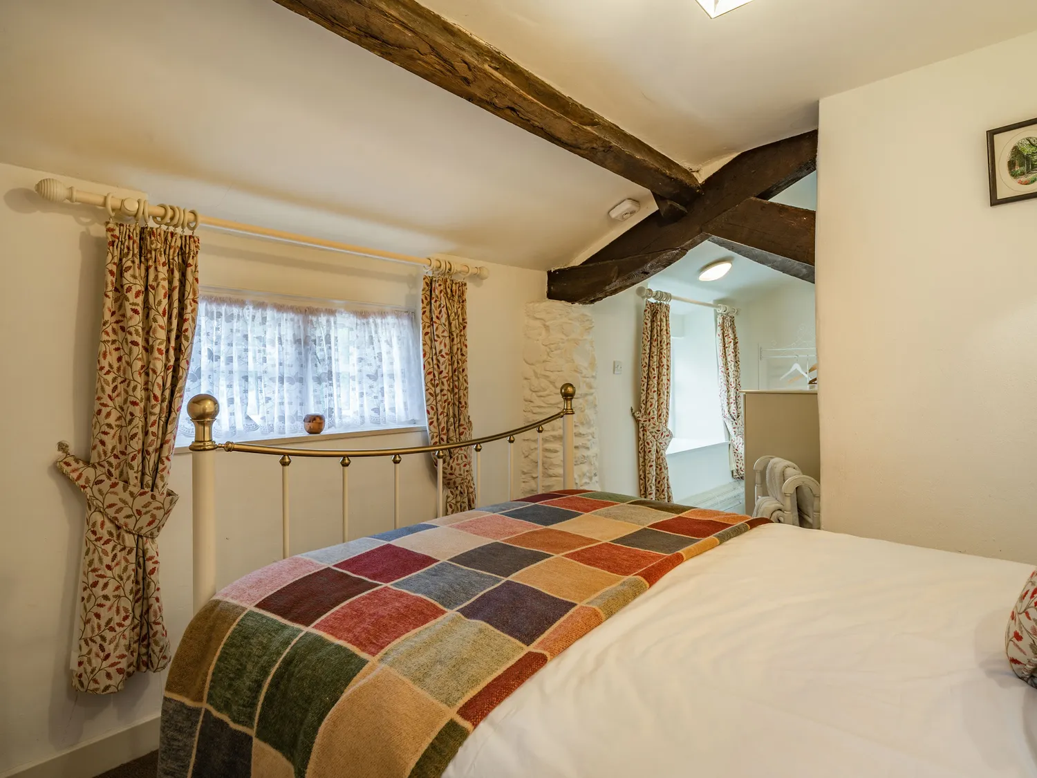 Cozy bedroom featuring a brass bed frame with a multicolored patchwork blanket, floral-patterned curtains, and exposed wooden beams, creating a charming and inviting atmosphere.