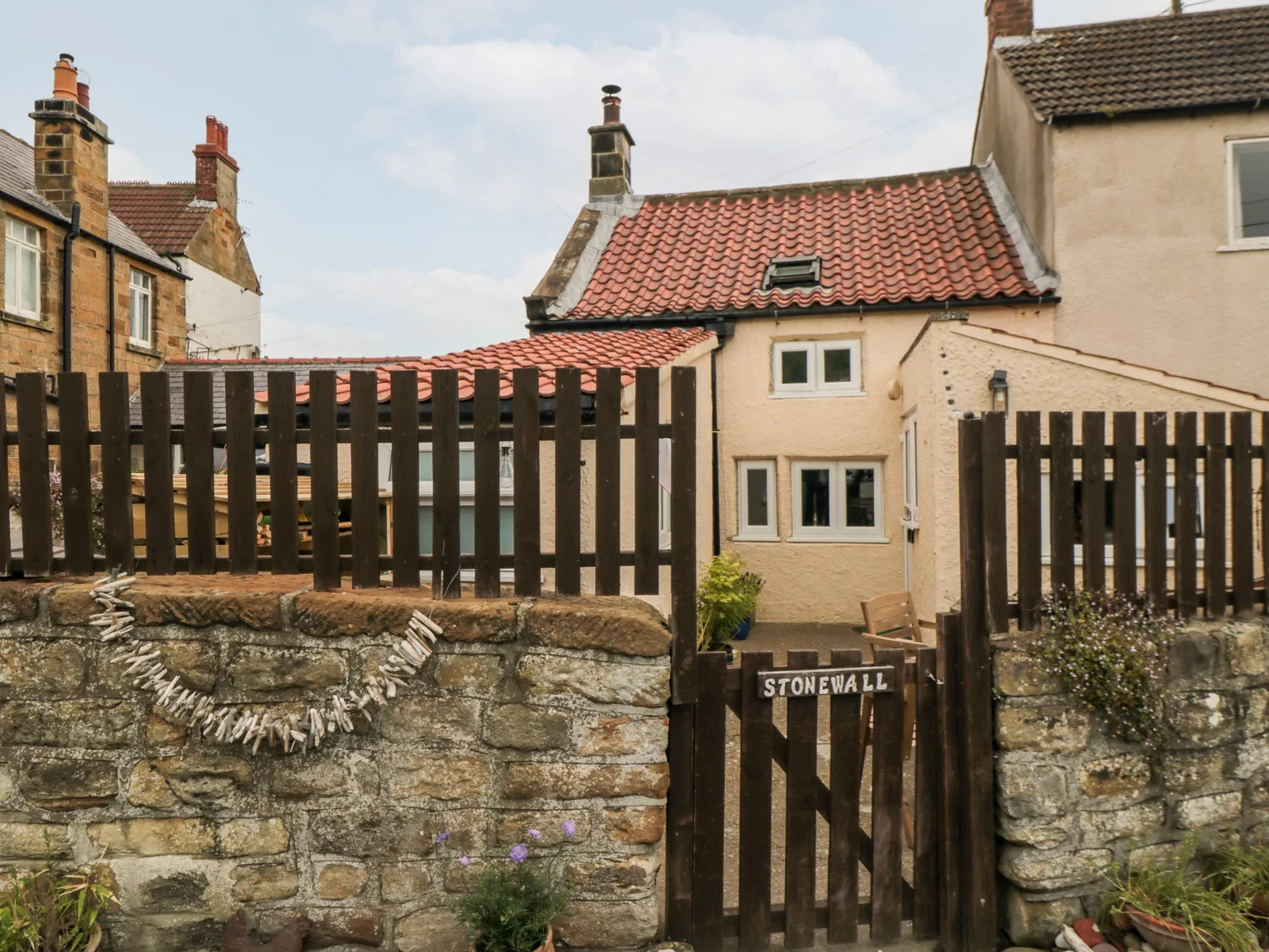 Charming cottage with red-tiled roof, wooden gate, and stone wall in a quaint village setting.