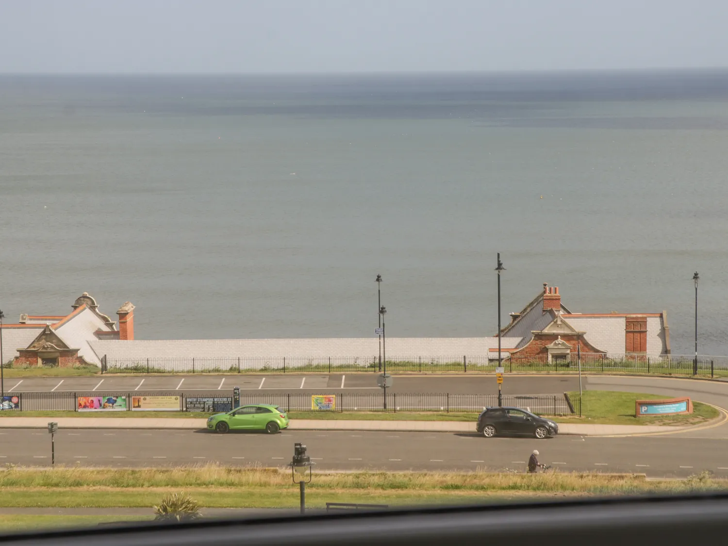 Scenic view of a coastal road with parked cars, featuring historic buildings and a calm sea in the background on a clear day.