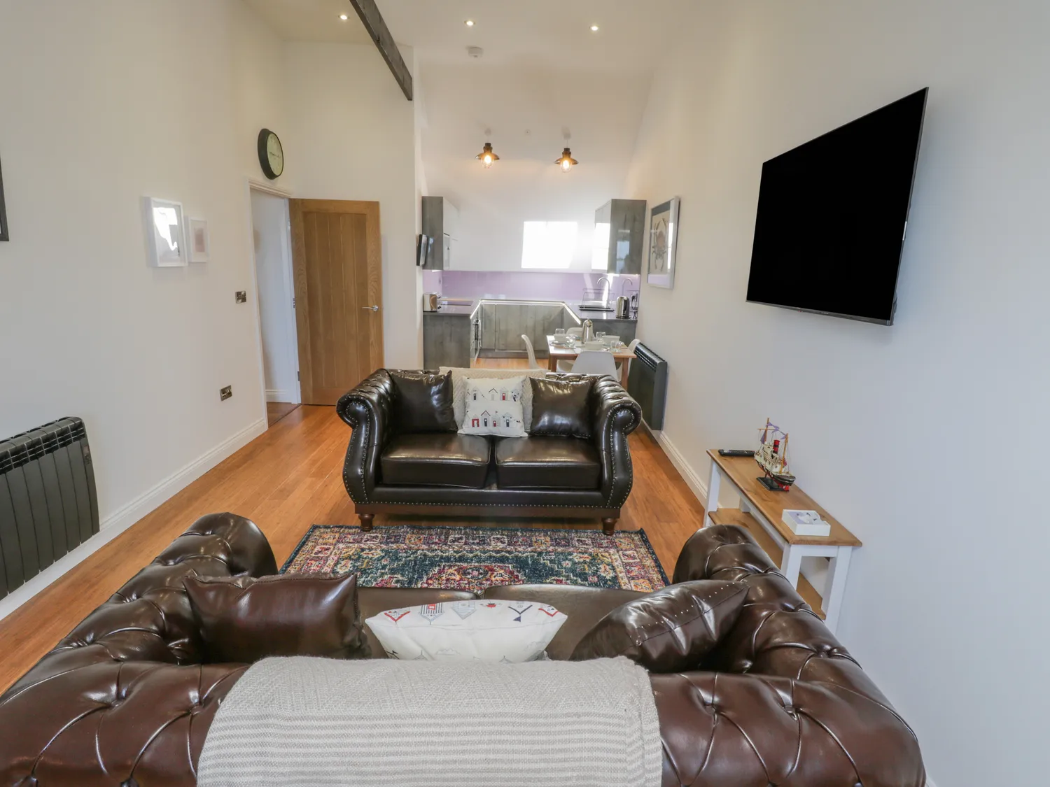 Modern living room featuring stylish leather sofas, a wooden coffee table, and a large flat-screen TV. The space has hardwood flooring, high ceilings with exposed beams, and an open kitchen area visible in the background. Natural light floods the room through large windows, creating a warm and inviting atmosphere.