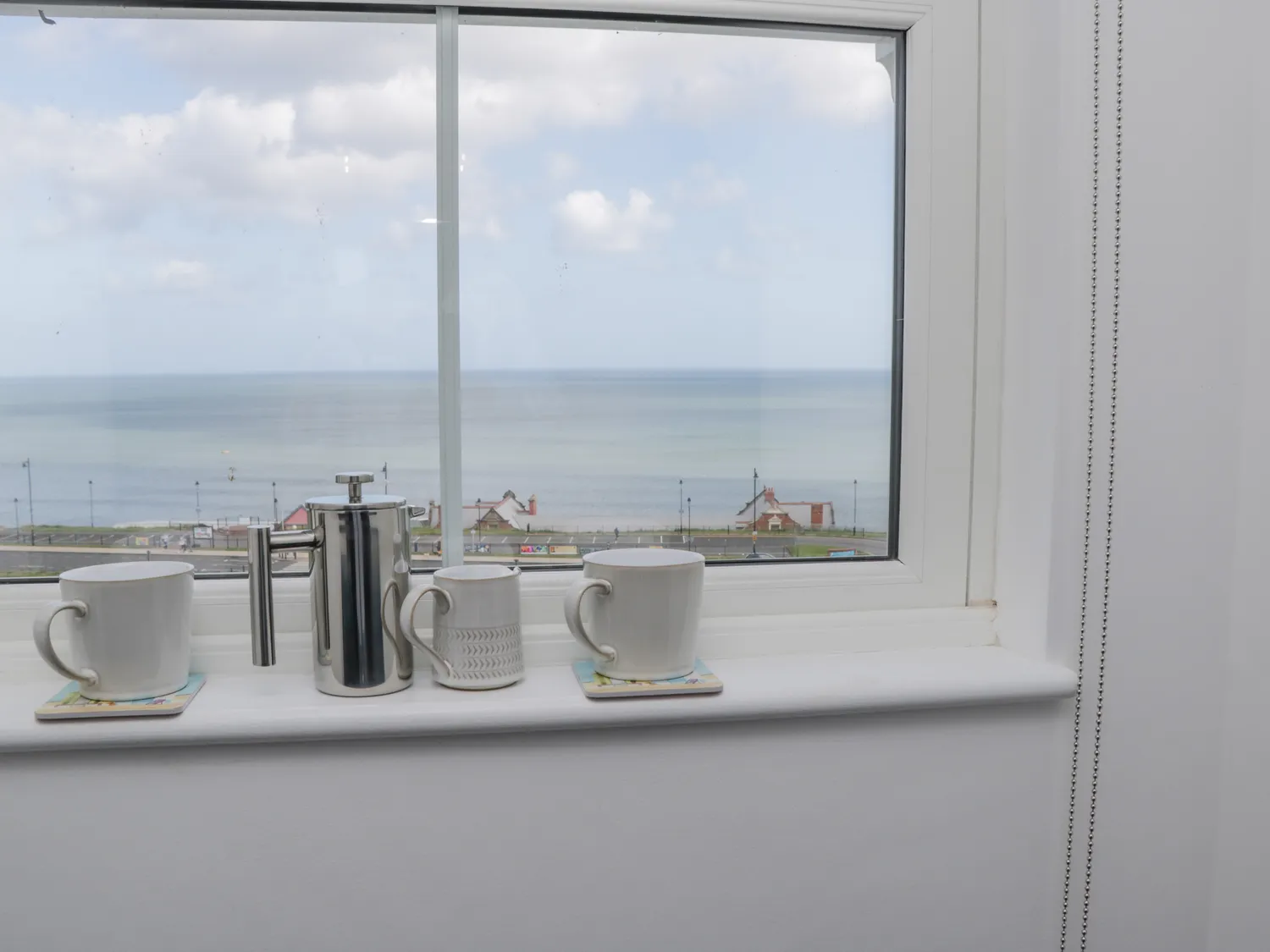 Cozy window view featuring two white mugs and a stainless steel French press on a windowsill, overlooking a serene seaside landscape with calm waters and distant buildings.