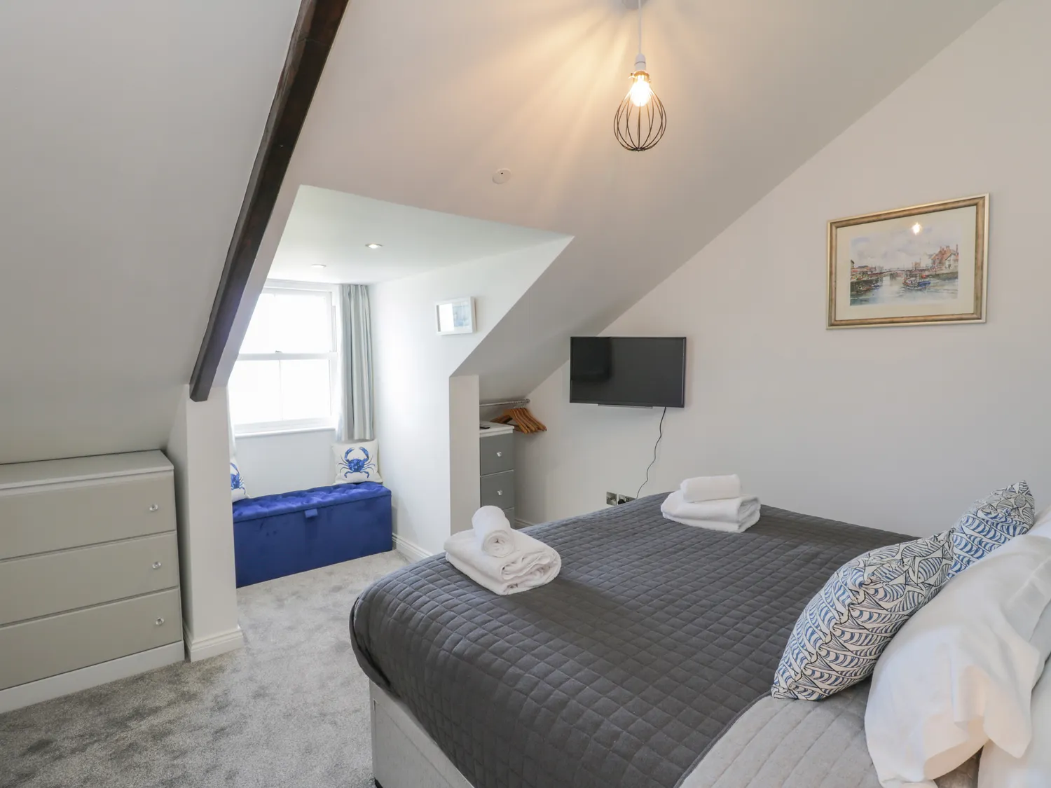 Cozy attic bedroom featuring a comfortable double bed with decorative pillows, a flat-screen TV, a storage dresser, and a blue bench by the window, illuminated by a stylish pendant light. Ideal for a relaxing stay.