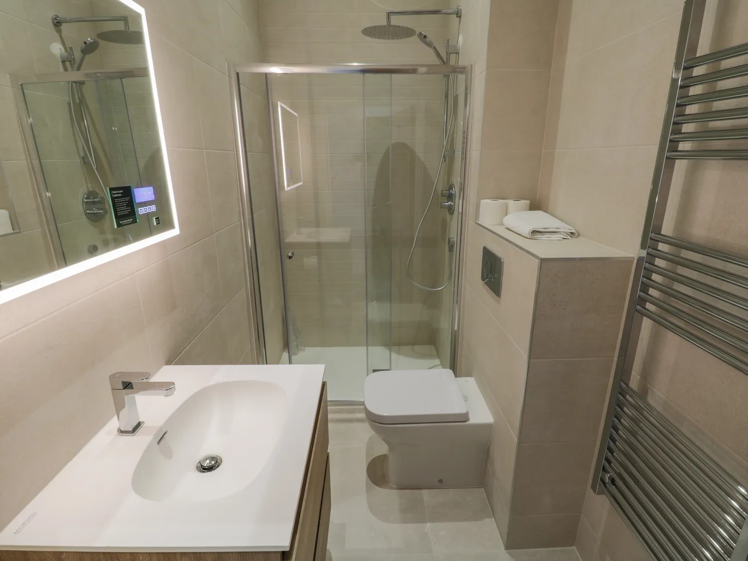 Modern bathroom featuring a glass shower enclosure, sleek vanity with a contemporary faucet, and a heated towel rail. The neutral color palette enhances the spacious feel, while the illuminated mirror adds a touch of elegance. Ideal for a stylish and functional home or hotel setting.