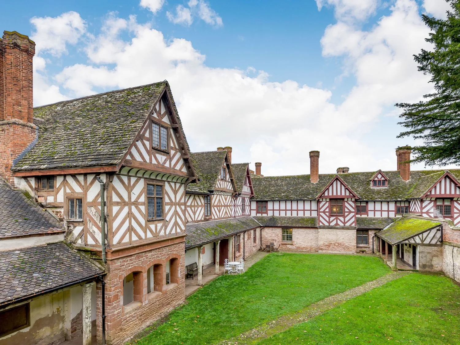 Historic Tudor-style building with timber framing and brick chimneys, featuring a green courtyard under a blue sky with scattered clouds.