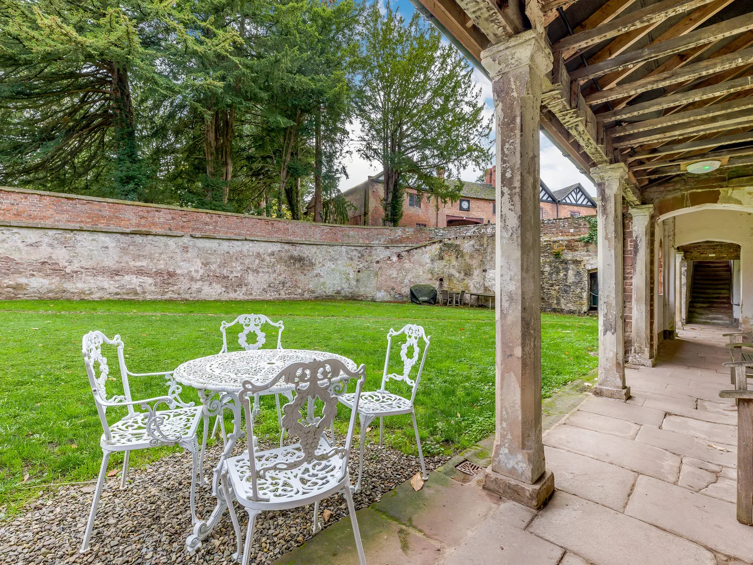 Outdoor garden with white wrought iron table and chairs, surrounded by lush greenery and historic brick walls, adjacent to a covered stone walkway.