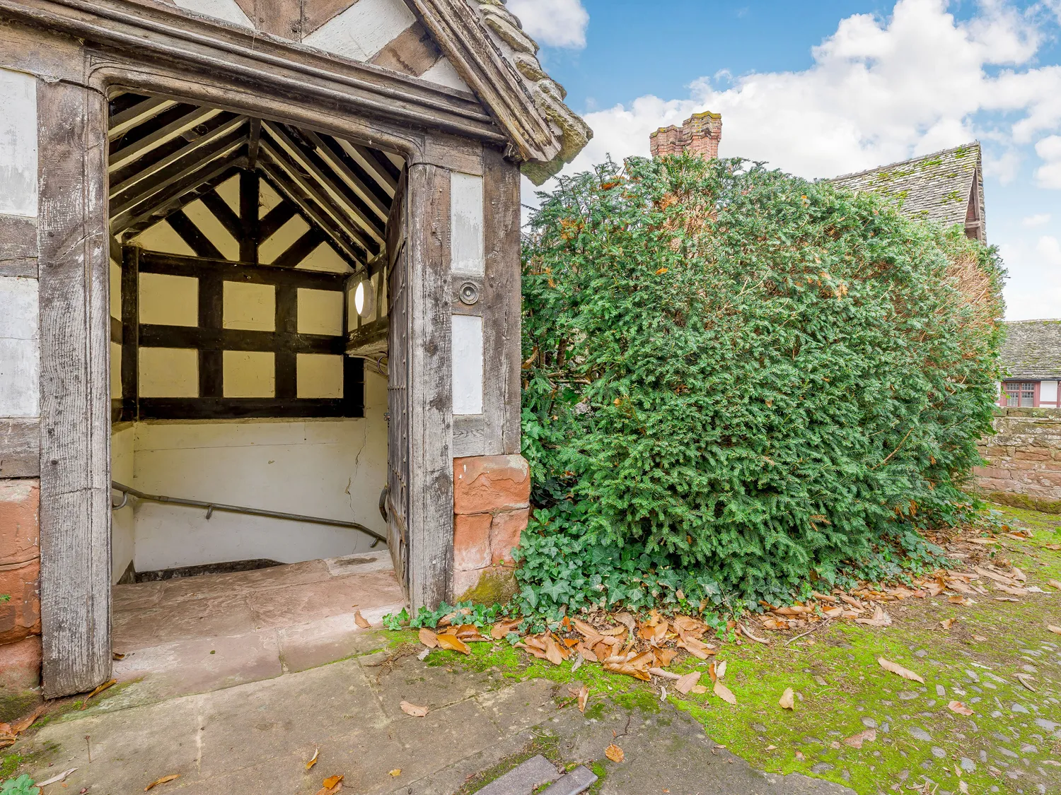 Entrance to a historic timber-framed building with a gabled roof, stone steps, and lush greenery in the background.