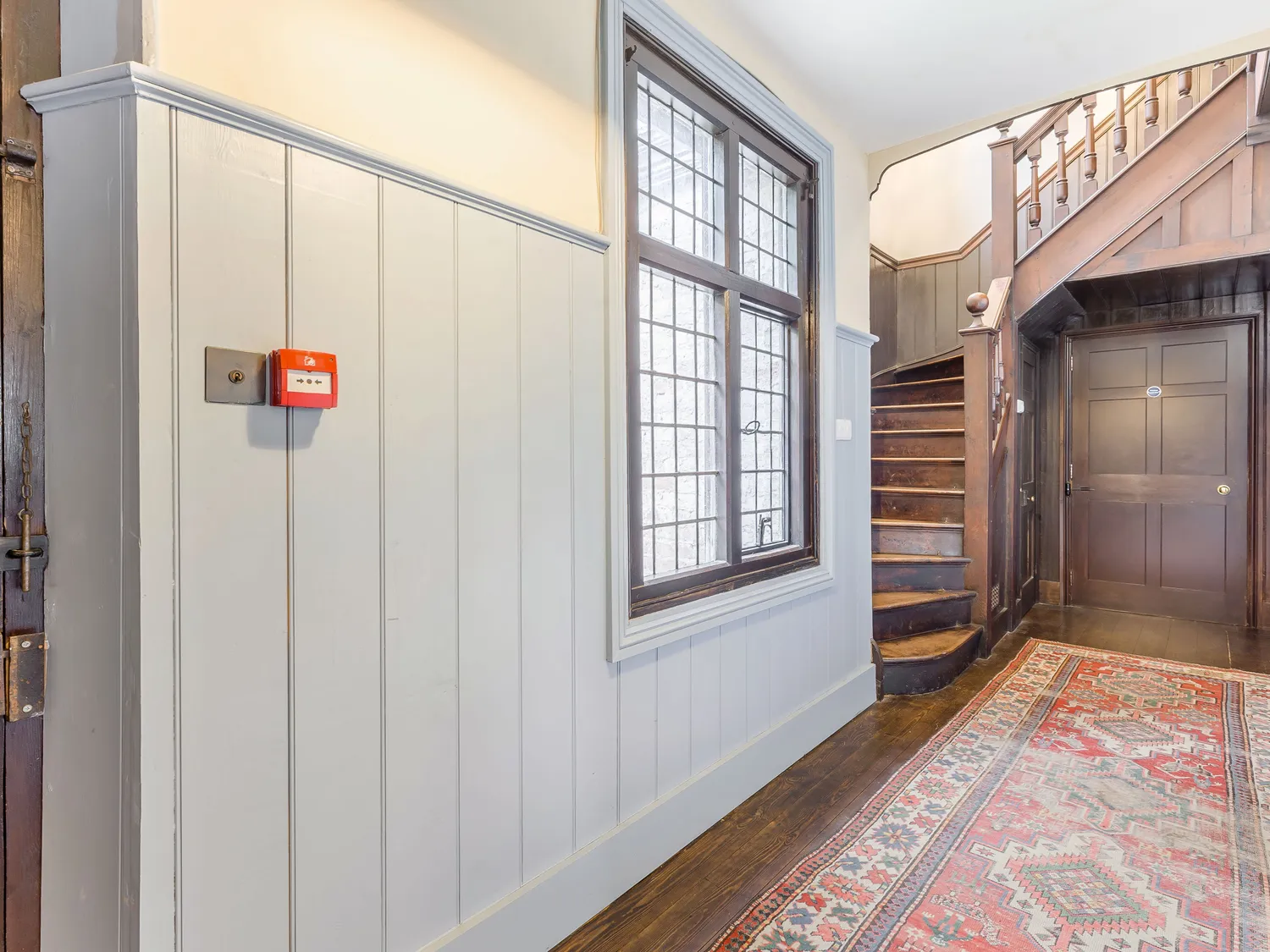 Traditional hallway with wooden staircase, vintage patterned rug, large window, and fire alarm on paneled wall.