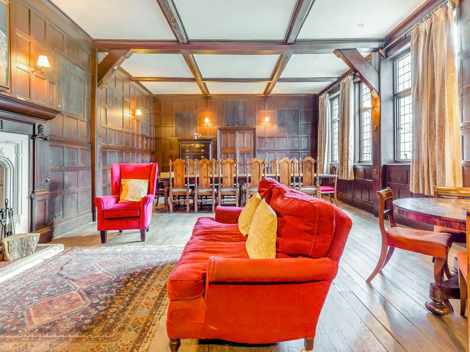 Historic dining room with wooden paneling, red armchairs, large dining table, and natural light from tall windows.