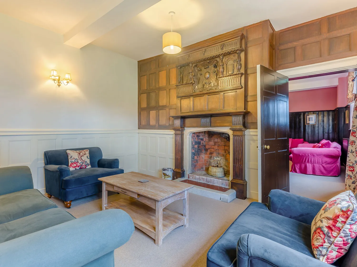 Cozy living room with vintage wooden fireplace, blue armchairs, and wooden coffee table, leading to a colorful adjoining room with pink furniture.