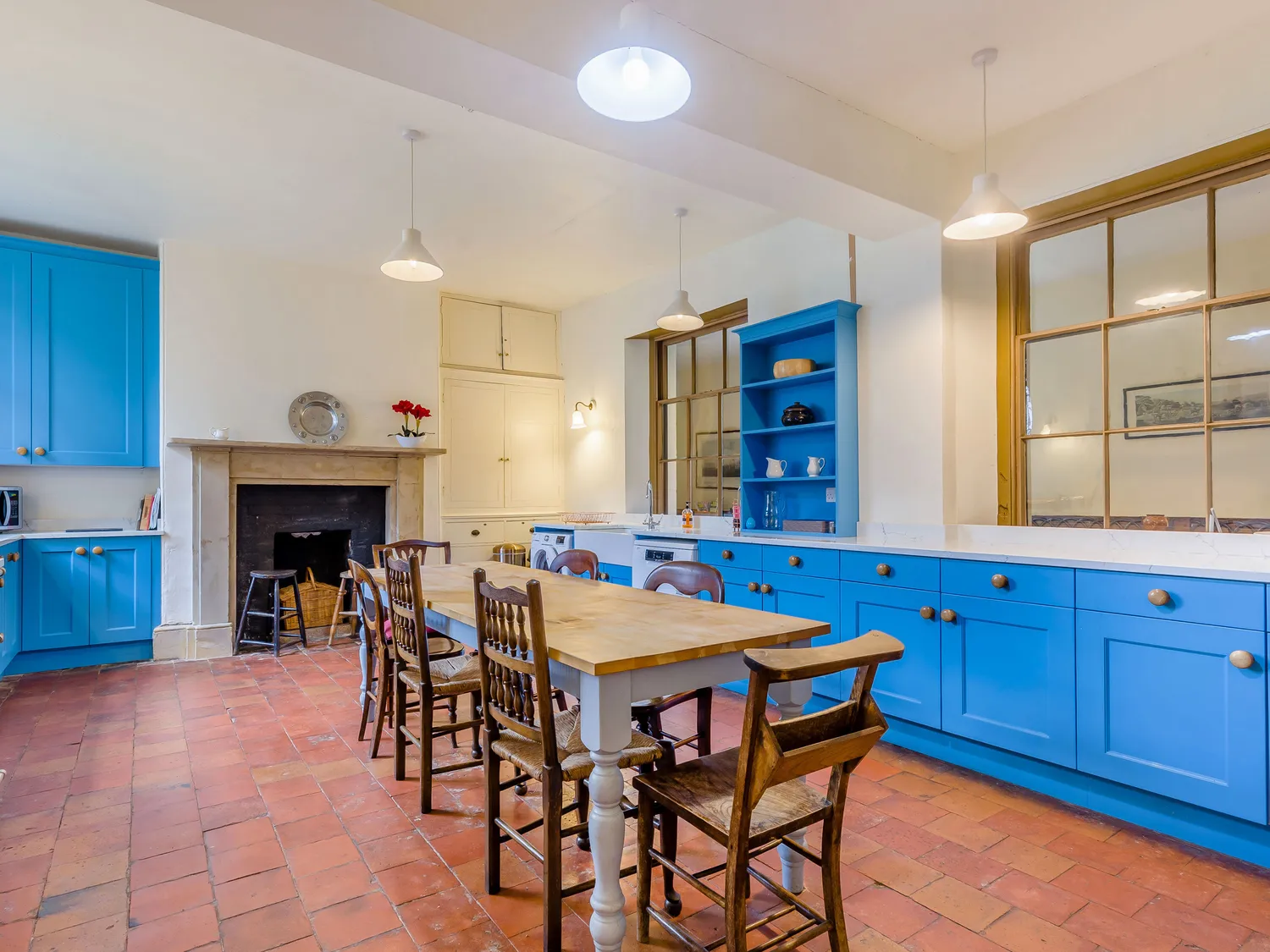 Spacious kitchen with blue cabinets, wooden dining table, vintage chairs, and terracotta tile flooring.