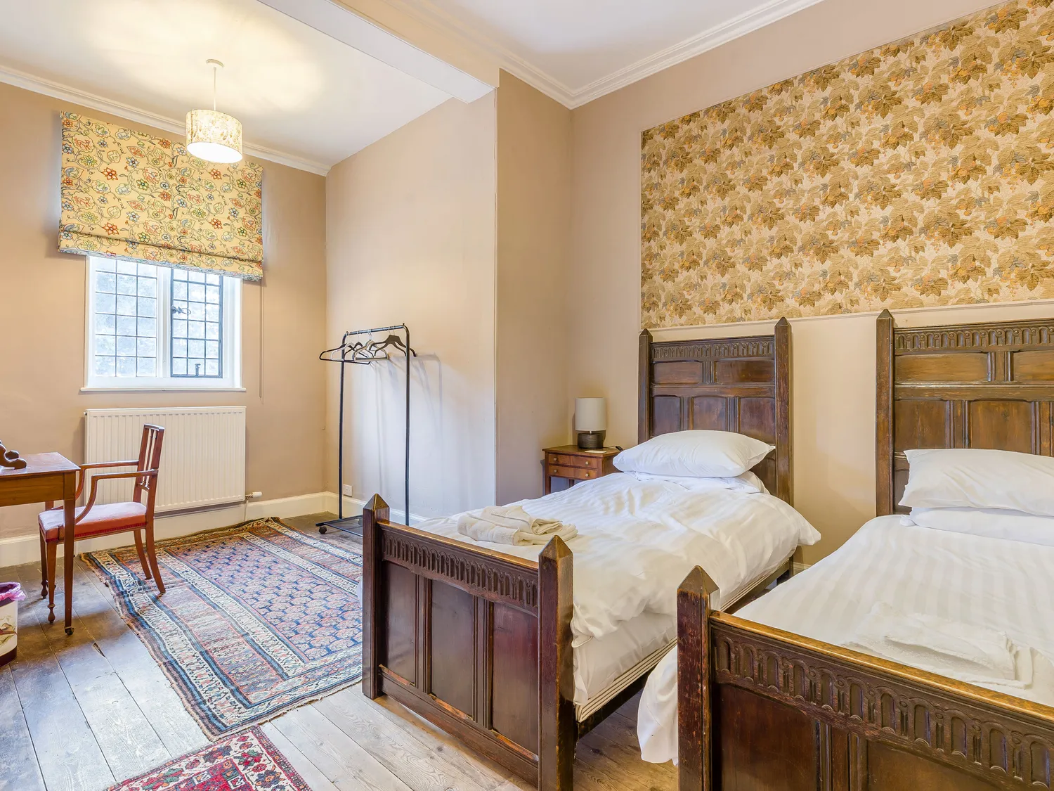 Cozy twin bedroom with vintage wooden beds, floral wallpaper, and a window with patterned blinds, featuring a small desk and chair, and a hanging clothes rack.