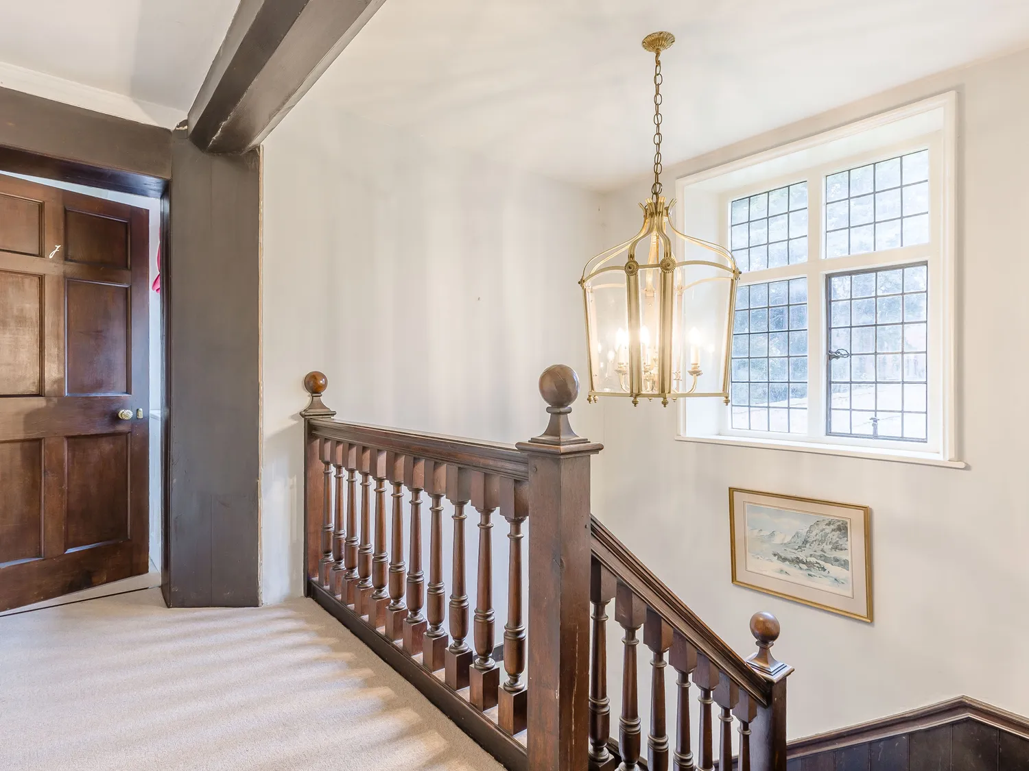 Traditional wooden staircase with ornate railing, large window with grid design, and elegant chandelier in a well-lit hallway.