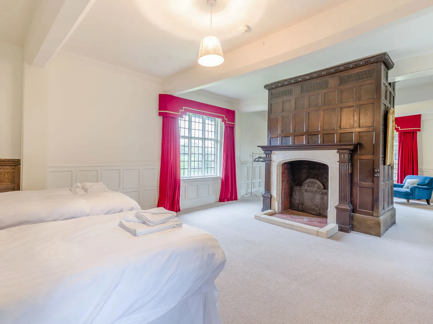 Spacious bedroom with twin beds, large wooden fireplace, red curtains, and natural light from a large window.