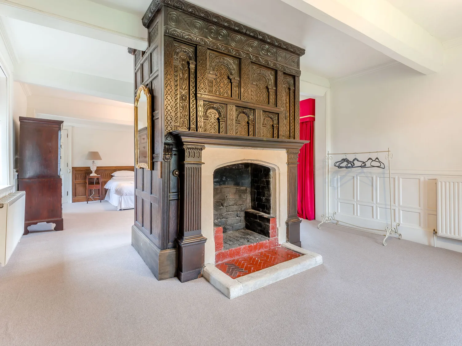 Spacious bedroom with ornate wooden fireplace, white walls, and vintage furniture.